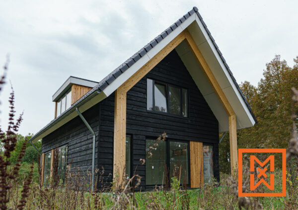 Moderne, zwart afgewerkte woning met grote ramen en zichtbare houten balken, gelegen in een veld met hoog gras. De woning heeft een strak, eigentijds ontwerp met een zadeldak en een dakkapel.