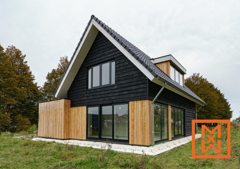 Moderne, zwarte en houten A-frame woning onder een bewolkte hemel. De woning heeft grote ramen en een strak design, een toonbeeld van hedendaagse architectuur. Het terrein is omgeven door een aangelegd gazon.