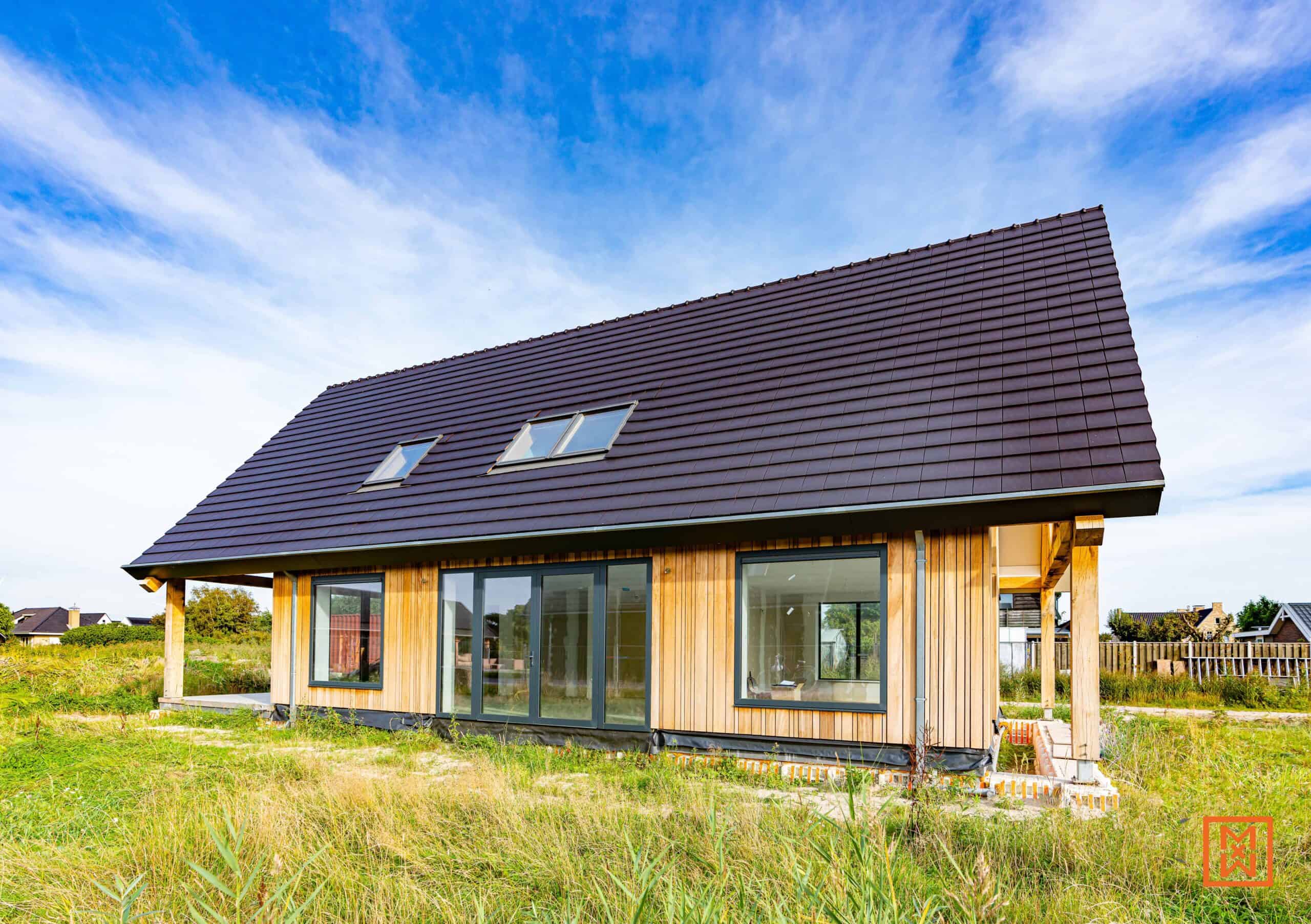 Moderne, gelijkvloerse woning met houten gevel, donkerbruin pannendak en grote ramen, gelegen in een grasveld onder een strakblauwe hemel. Drie dakramen zijn zichtbaar op het dak. De woning is een toonbeeld van eigentijdse architectuur en duurzaam design.