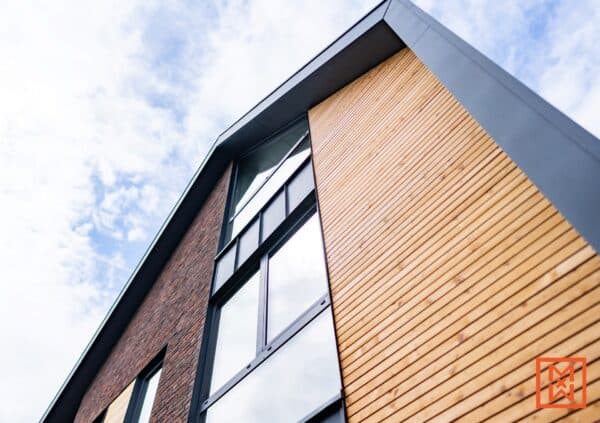 Van onderaf gezien de buitenkant van een modern gebouw, met een opvallende combinatie van baksteen en lichte houten gevelbekleding. Grote ramen met zwarte kozijnen zijn prominent aanwezig in het bakstenen gedeelte, terwijl het houten gedeelte strakke, horizontale lijnen heeft. De lucht is erboven zichtbaar.