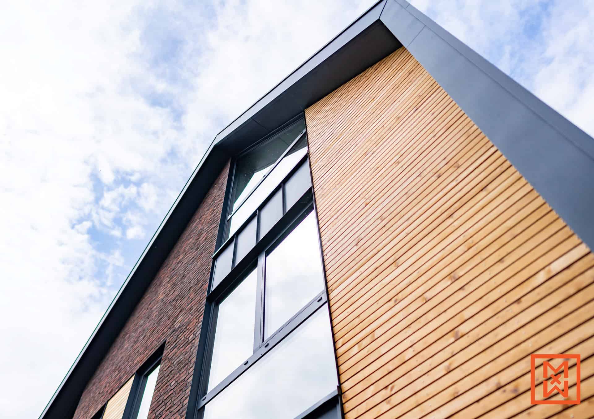 Van onderaf gezien de buitenkant van een modern gebouw, met een opvallende combinatie van baksteen en lichte houten gevelbekleding. Grote ramen met zwarte kozijnen zijn prominent aanwezig in het bakstenen gedeelte, terwijl het houten gedeelte strakke, horizontale lijnen heeft. De lucht is erboven zichtbaar.