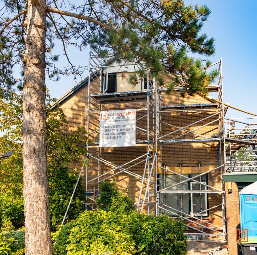 Een bakstenen huis wordt gerenoveerd, omgeven door steigers en weelderig groen. Een spandoek van "Van Weenen Bouwbedrijf" adverteert nieuwbouw, renovatie, uitbreidingen en raaminstallaties. Het zonnige tafereel toont woningverbetering en bouwwerkzaamheden in uitvoering.