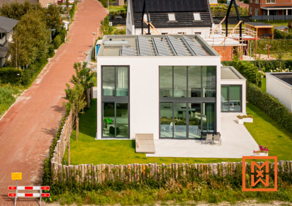 Luchtfoto van een modern, wit minimalistisch huis met zonnepanelen op het dak, gelegen op een grasveld met een houten schutting. Het huis heeft grote glazen ramen en een terras met tuinmeubilair. Op de achtergrond is een pad van rode baksteen zichtbaar.