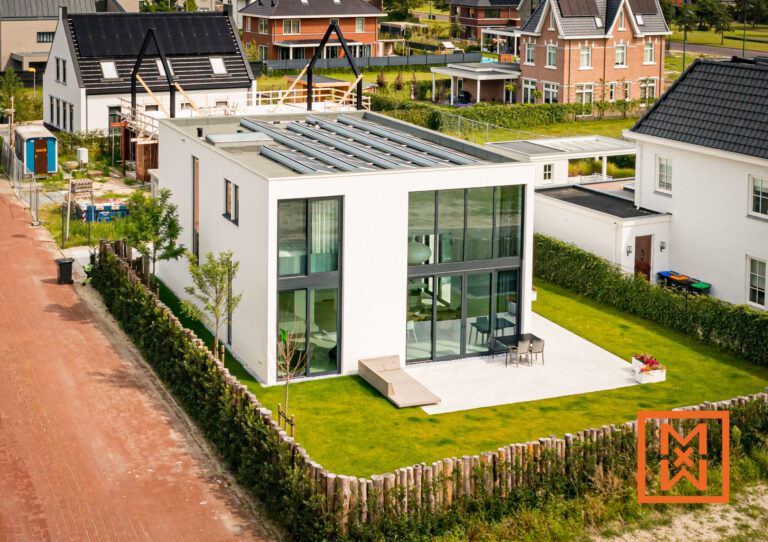 Luchtfoto van een modern, wit huis met twee verdiepingen, een plat dak en zonnepanelen. Het huis heeft grote, donkere ramen en een minimalistisch design. Het staat op een verzorgd gazon met een houten schutting en tuinmeubilair. Nabijgelegen huizen en een pad van rode baksteen zijn zichtbaar.