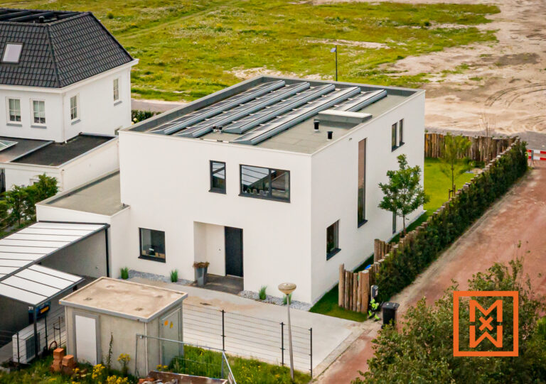 Luchtfoto van een moderne, witte woning met twee verdiepingen en een plat dak met zonnepanelen. De woning heeft een minimalistisch ontwerp met donkere kozijnen en een grijze voordeur. Een kleine, betegelde oprit en een keurig aangelegde tuin omringen de woning, gelegen in een nieuwbouwwijk.