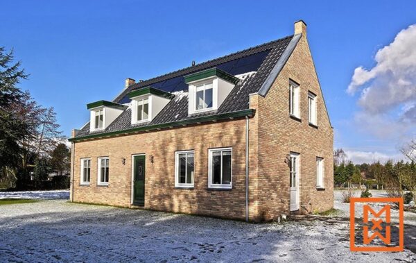 Een charmant, lichtbruin bakstenen huis met een donkergrijs pannendak en zonnepanelen staat op een besneeuwd gazon. Dakkapellen met donkergroene biezen zorgen voor een architectonisch detail. Het zonnige wintertafereel suggereert een vredige, idyllische omgeving.