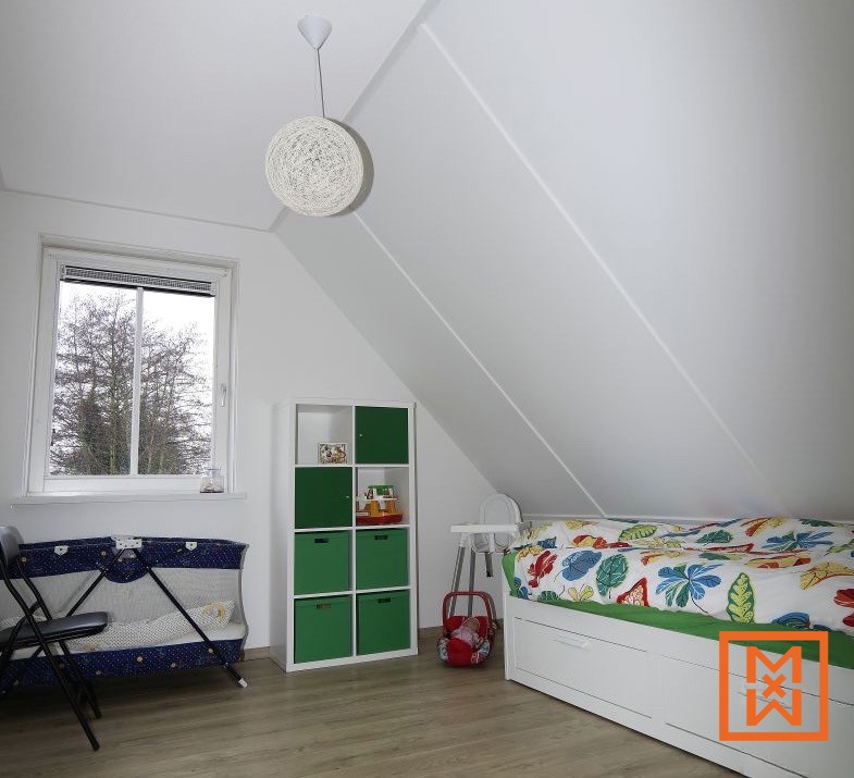 Lichte, luchtige zolderkamer met schuine plafonds, een wit uitschuifbed met kleurrijk bladerdekbed, een groene opbergkubus en een babybedje. Natuurlijk licht stroomt binnen via een raam met uitzicht op de bomen.