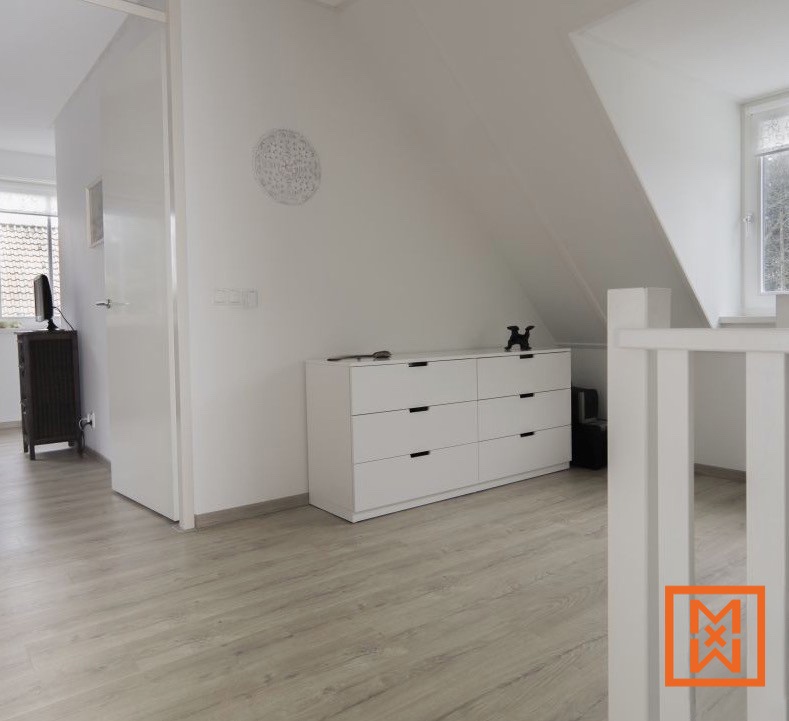 Een moderne witte ladekast met zes lades staat in een lichte, minimalistische zolderkamer. De lichtgrijze vloer met houtlook en de witte muren zorgen voor een strakke, ruimtelijke sfeer. Er staat een klein zwart decoratief object bovenop.