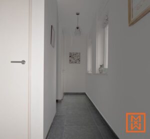 Lichte, minimalistische hal met donkergrijze tegelvloer, witte muren en inbouwspots. Een collage van foto's en een hanglamp in een kooi geven de ruimte karakter. De hal heeft een witte deur en raam met witte gordijnen.