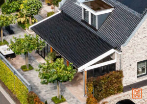 Strakke, moderne woning met donker dak en lichte stenen gevel. Sierlijke bomen en een groene haag omlijsten het pad naar de overdekte entree.