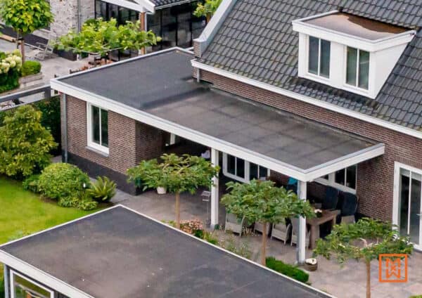 Bovenaanzicht van een modern huis met plat dak en een overdekt terras met tuinmeubelen. De bakstenen gevel en de groene tuin creëren een rustige sfeer.