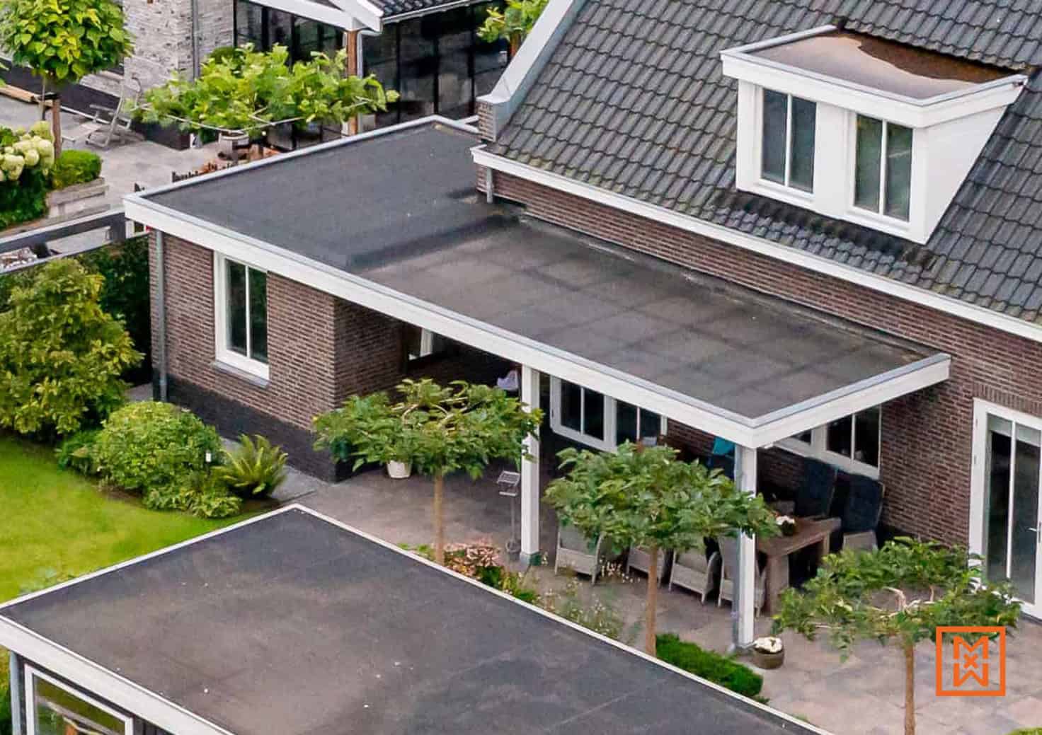Bovenaanzicht van een modern huis met plat dak en een overdekt terras met tuinmeubelen. De bakstenen gevel en de groene tuin creëren een rustige sfeer.