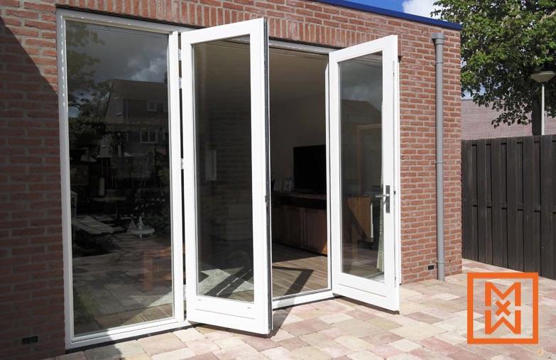 Witte openslaande terrasdeuren geven toegang tot een stenen terras met uitzicht op een woonkamer met een flatscreen-tv. De deuren maken deel uit van een modern huis met een rode bakstenen buitenkant en een donkerhouten hek op de achtergrond. Het zonlicht stroomt naar binnen en creëert een lichte en uitnodigende sfeer.