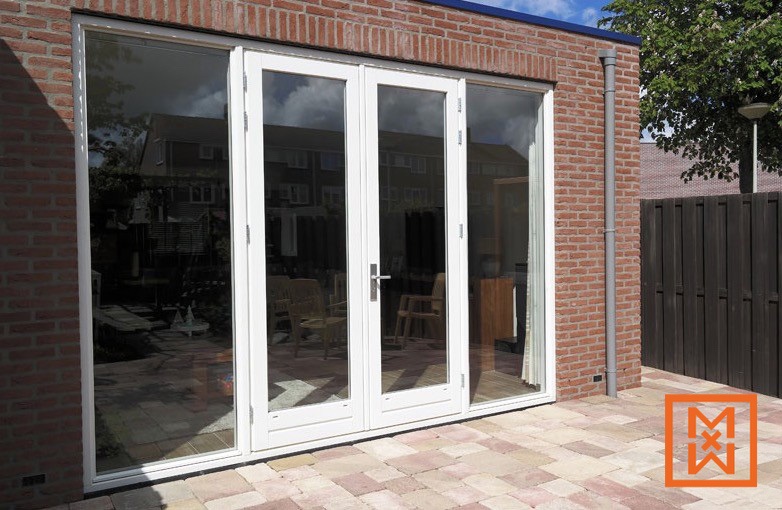 Witte openslaande deuren, onderdeel van een schuifpuisysteem, geven toegang tot een stenen terras. De deuren zijn in een rode bakstenen muur geplaatst, met grote ramen ernaast, die een zonnig uitzicht op de achtertuin bieden. De algehele uitstraling is modern en strak.