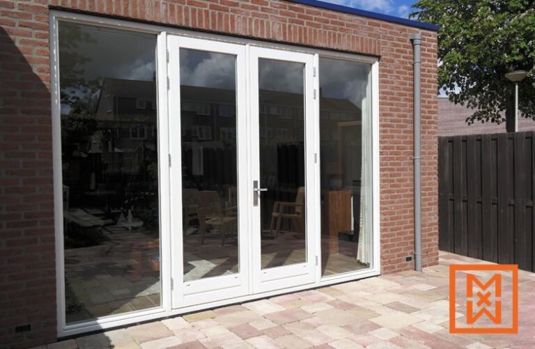 Witte openslaande deuren met glazen panelen geven toegang tot een stenen terras met uitzicht op de achtertuin. De deuren maken deel uit van een moderne uitbouw van een bakstenen huis.