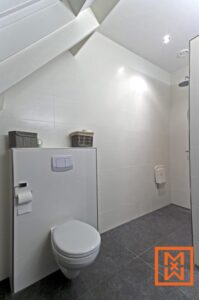 Moderne, minimalistische badkamer met witte wandtegels, een wandcloset en een inloopdouche. De donkergrijze vloertegels contrasteren scherp met de helderwitte armaturen. Een plankje boven het toilet biedt opbergmandjes.
