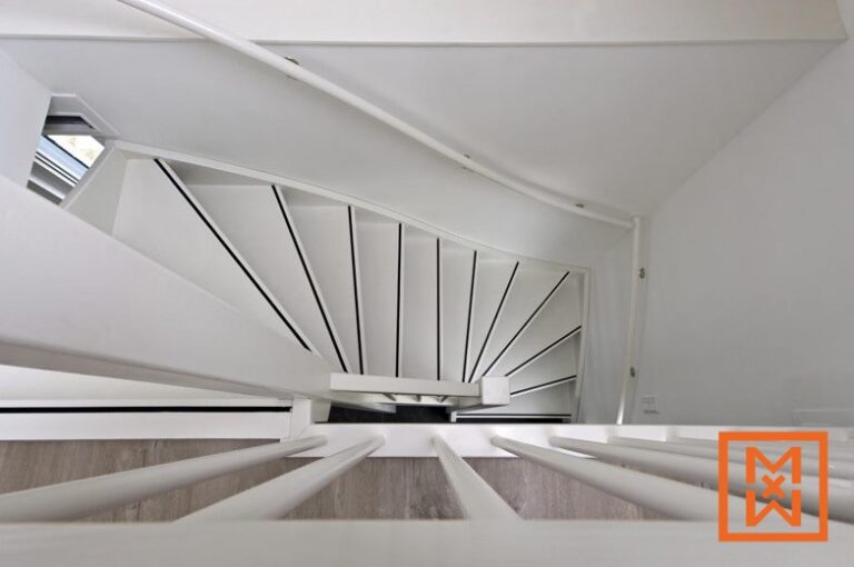 Bovenaan een moderne, witte wenteltrap, van onderaf gezien, zijn de slanke, met zwart geaccentueerde treden te zien die omhoog slingeren. De trap heeft een minimalistische witte leuning.