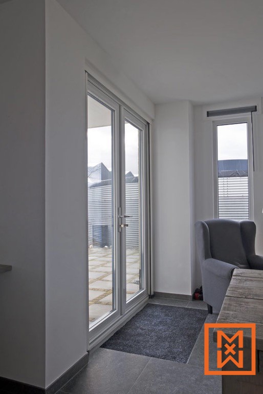 Moderne aluminium openslaande deuren geven toegang tot een terras met een stenen vloer, met uitzicht op een gegolfd metalen hek en de huizen daarachter. Een donkergrijze fauteuil en houten tafel zijn zichtbaar in de hoek van de kamer, wat zorgt voor een minimalistisch, licht interieur.