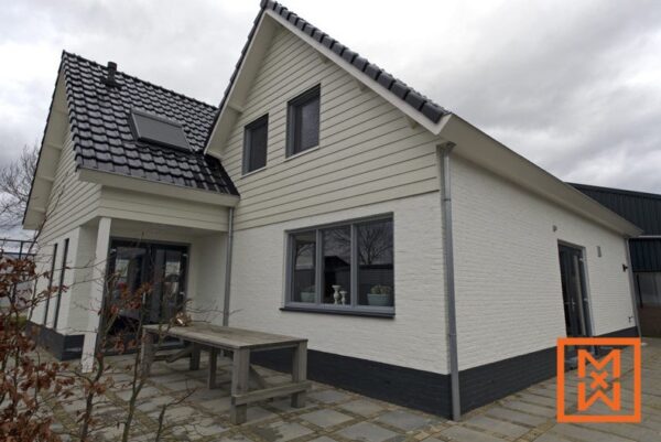 Moderne buitenkant van het huis met lichtbeige houten gevelbekleding en strak witte bakstenen. Donkergrijze kozijnen en een donkere plint completeren het ontwerp. Op de voorgrond is een terras met stenen tegels en een houten tafel zichtbaar.