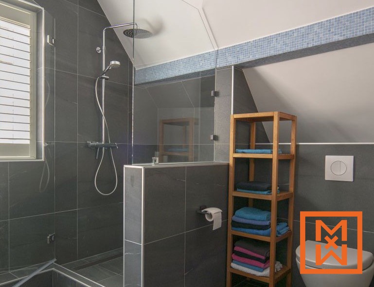 Moderne badkamer met grijze tegels, een inloopdouche, een houten plankje met netjes opgevouwen handdoeken en een toilet. De douche is voorzien van een regendouche en een glazen omkasting. Een kleine rand van blauwe mozaïektegels accentueert het schuine plafond.