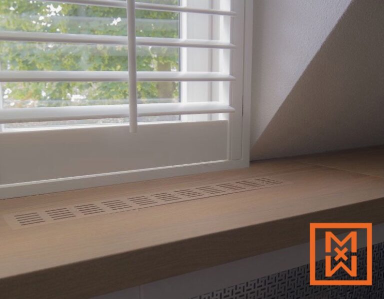 Close-up van een lichte houten vensterbank met een discreet ventilatierooster, onder witte luiken in een raam met uitzicht op weelderige groene bomen. De vensterbank maakt deel uit van een stijlvolle, moderne raaminstallatie.