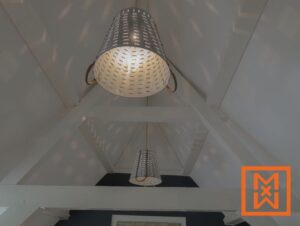 Drie gegalvaniseerde metalen lampen hangen aan een wit, schuin plafond met zichtbare balken en werpen zacht licht. De verlichting in rustieke boerderijstijl creëert een warme en uitnodigende sfeer.
