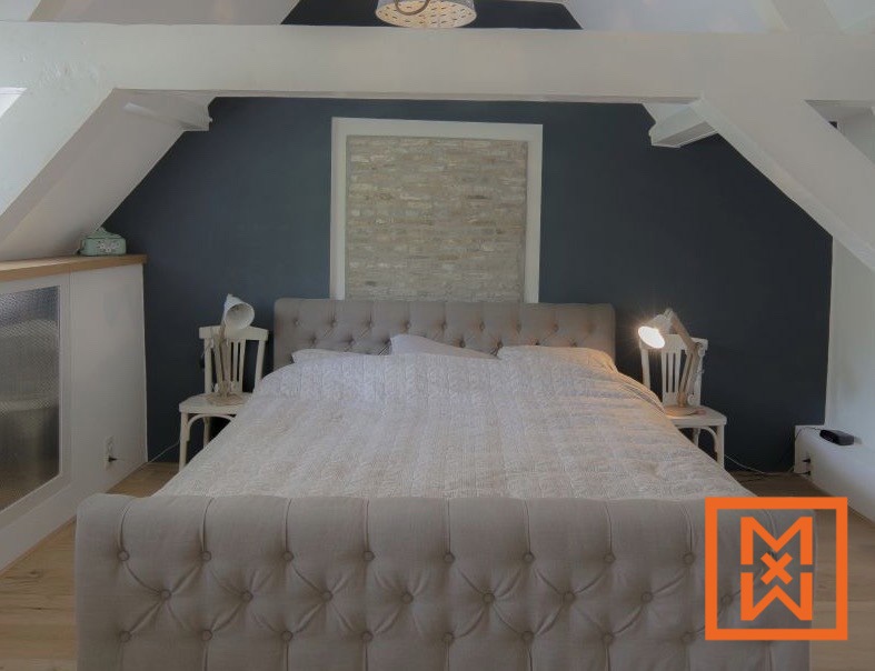 Beige bed met gecapitonneerd hoofdeinde in een gezellige zolderkamer met donkergrijze muren en een lichte stenen accentmuur. Twee bedlampjes flankeren het bed en dragen bij aan de rustige en uitnodigende sfeer.