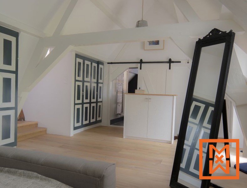 Lichte, luchtige zolderslaapkamer met een licht eikenhouten vloer, ingebouwde donkergrijze en witte paneelkasten en een grote zwarte spiegel. Een moderne schuurdeur scheidt de slaapkamer van een kleinere kamer.
