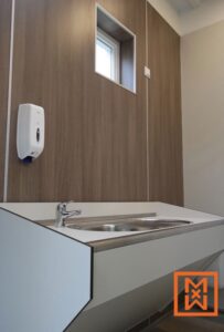 Moderne, witte wandwastafel met roestvrijstalen kom en chromen kraan in een strak, licht toilet. Een witte zeepdispenser is bevestigd aan de met hout beklede wand. Een klein raam zorgt voor natuurlijk licht.