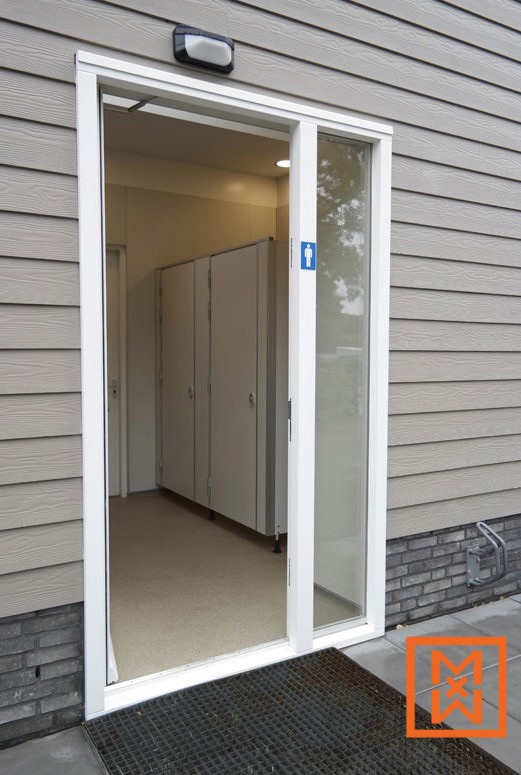 Moderne, toegankelijke toiletingang met een glazen schuifdeur en grijze gevelbekleding. Binnenin zijn strakke, lichtgrijze hokjes te zien. Op de deur is een symbool van een herentoilet zichtbaar.