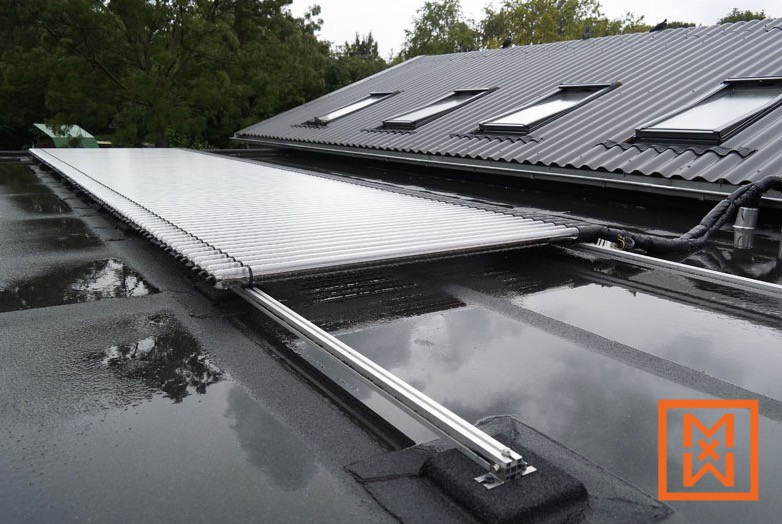 Een lang, plat zonnecollectorsysteem is gemonteerd op een donker, nat dak. Het systeem bestaat uit talloze transparante buizen en is bevestigd aan een aluminium rail. Op de achtergrond is een donkergrijs zadeldak met diverse dakramen te zien. Regenwater is zichtbaar op het dakoppervlak.
