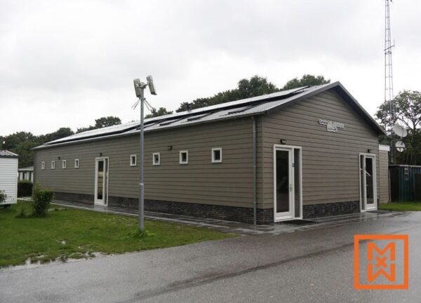Modern, grijs modulair gebouw met zonnepanelen op het dak. Het gebouw heeft meerdere ramen en twee dubbele deuren en is gelegen in een grasveld vlakbij andere gebouwen.