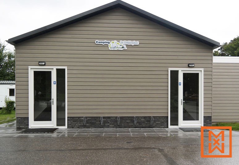 Modern, beige kampeertoiletgebouw met witte deuren en de tekst "Camping De Keienplant". Het gebouw heeft een donkergrijs dak en staat op een verhard terrein met fietsenrekken.