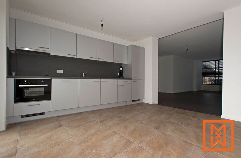 Moderne, lichtgrijze keuken met donkergrijs aanrechtblad en ingebouwde oven, die uitkomt op een ruime woonkamer met donkere hardhouten vloeren en een groot raam. De algehele stijl is minimalistisch en eigentijds.