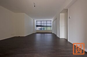Lege, moderne woonkamer met donkere hardhouten vloeren en een groot raam met zwarte jaloezieën. De muren zijn wit geverfd, wat zorgt voor een licht en ruimtelijk gevoel. Er hangt een enkele gloeilamp aan het plafond.