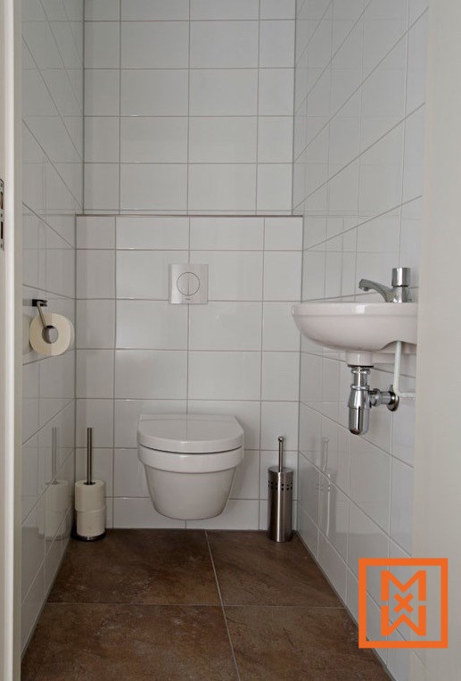 Kleine, moderne badkamer met witte tegels, een hangend toilet, een kleine wastafel en bruine vloertegels. Toiletpapier en een toiletborstel zijn netjes geplaatst. Het minimalistische ontwerp zorgt voor een strakke en efficiënte ruimte.
