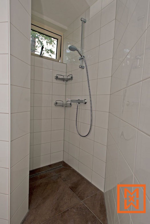 Moderne inloopdouche met witte tegelwanden, bruine vloertegels en een chromen douchekop en -armaturen. Twee kleine chromen planchetten zijn aan de muur bevestigd. Een klein raam zorgt voor natuurlijk licht.
