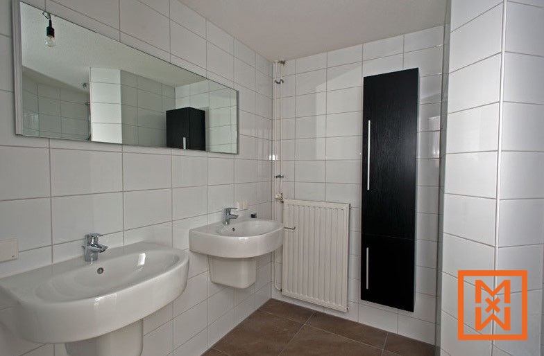 Moderne, wit betegelde badkamer met twee wastafels aan de wand, een zwarte handdoekradiator en een rechthoekige spiegel erboven. De vloer is betegeld met donkerbruine tegels.