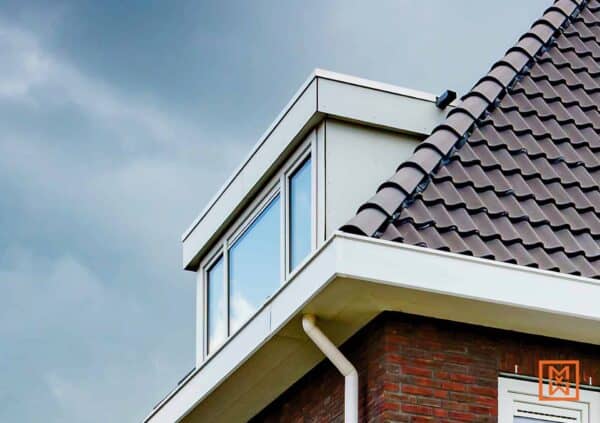 Detail van een huis met een dakraam, bruine dakpannen, witte dakranden en een rode bakstenen muur tegen een bewolkte lucht.