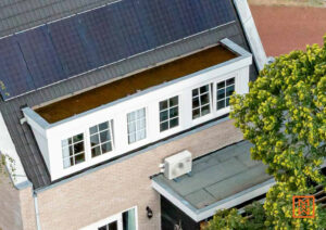 Bovenaanzicht van een huis met zonnepanelen en een dakkapel met witte kozijnen en ramen met roeden. Een airco-unit staat op het platte dak van een aanbouw.