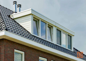 Dakkapel met witte kozijnen en vijf ramen in een donker pannendak boven een bakstenen muur. Twee metalen schoorstenen steken boven de dakkapel uit. Heldere hemel op de achtergrond.