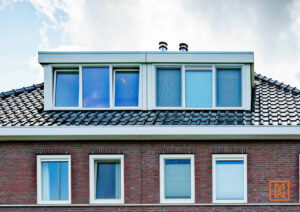 Een stenen huis met rode bakstenen en witte kozijnen. Boven ramen onder een dak met zwarte dakpannen. Twee schoorstenen steken boven het dak uit.