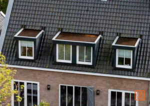 Dakraam met drie dakkapellen op een donker dak van een bakstenen huis. Witte ramen contrasteren met de donkere dakpannen.
