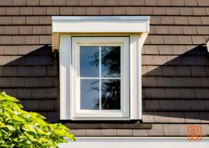 Een wit raam met roedeverdeling in een donkerbruin dak met dakpannen. Het raam heeft een witte omlijsting en een witte kroonlijst erboven.