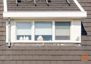 Raam met drie ruiten in een dak, omgeven door grijze dakpannen. Witte kozijnen en een witte omlijsting geven een strakke uitstraling. Binnen staan vazen.