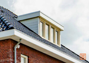 Dakraam op een huis met rode bakstenen en donkere dakpannen. Het dakraam heeft een lichtgrijze omlijsting en houten kozijnen. Een witte dakgoot loopt langs het dak.