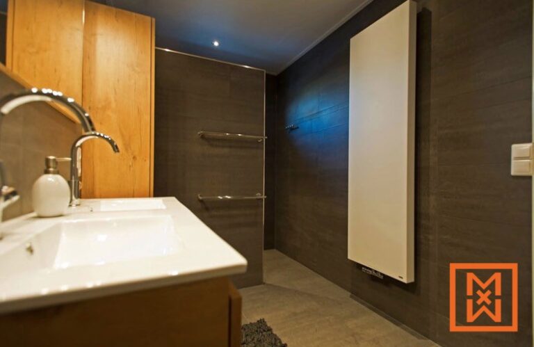 Moderne badkamer met een strakke, witte handdoekradiator, een wastafel met dubbele wastafel, lichthouten kasten en donkerbruine tegelwanden. De algehele esthetiek is minimalistisch en verfijnd.