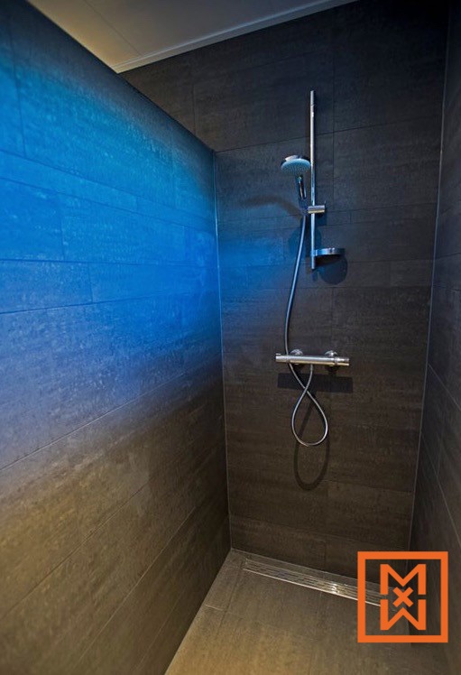 Moderne inloopdouche met donkergrijze tegels en een strak chromen douchearmatuur. Eén wand is voorzien van blauwe accentverlichting, wat een rustgevende spa-achtige sfeer creëert.