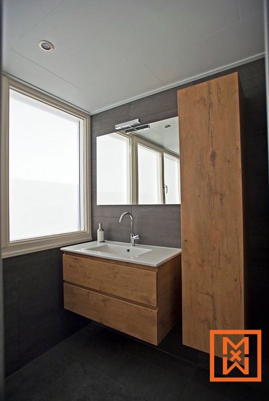 Moderne badkamer met een zwevend wastafelmeubel van licht hout en een hoge, bijpassende opbergkast. Een strakke witte wastafel en chromen kraan staan boven het wastafelmeubel, tegen een donkergrijze betegelde muur. Natuurlijk licht stroomt binnen via een matglasraam.
