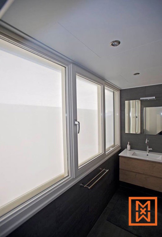 Moderne badkamer met een strak schuifraam met drie ruiten en matglas voor privacy. De donkergrijze betegelde muren en het lichte houten wastafelmeubel zorgen voor een verfijnd contrast. Onder het raam is een handdoekhouder gemonteerd.